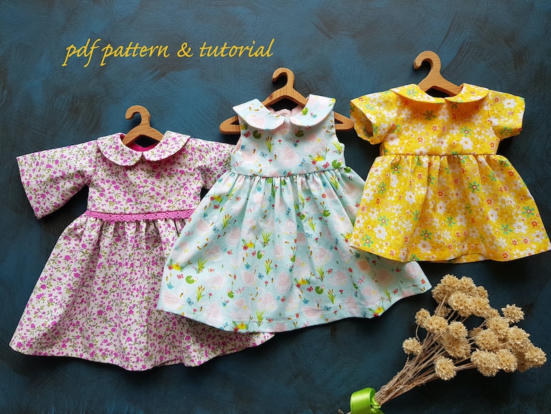 Puede incluir: Tres vestidos de mu&ntilde;eca en perchas, cada uno con un estampado floral diferente. Los vestidos tienen un cuello Peter Pan y son sin mangas, de manga corta y de manga larga. El texto "pdf pattern & tutorial" es visible en la parte superior de la imagen.