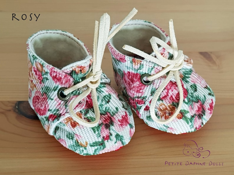 May include: A pair of white fabric baby shoes with a floral print and cream-colored laces. The shoes have a red trim and are labeled "Rosy".
