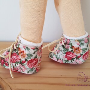 May include: A pair of doll shoes with white laces and a floral print. The shoes are made of a soft, pink and green fabric with a white trim. The shoes are tied with white laces and have a red sole.