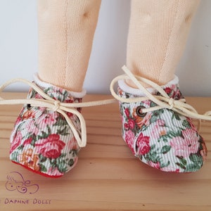 May include: A pair of floral print doll shoes with white laces and red trim. The shoes are tied with a bow and have a white trim around the top.