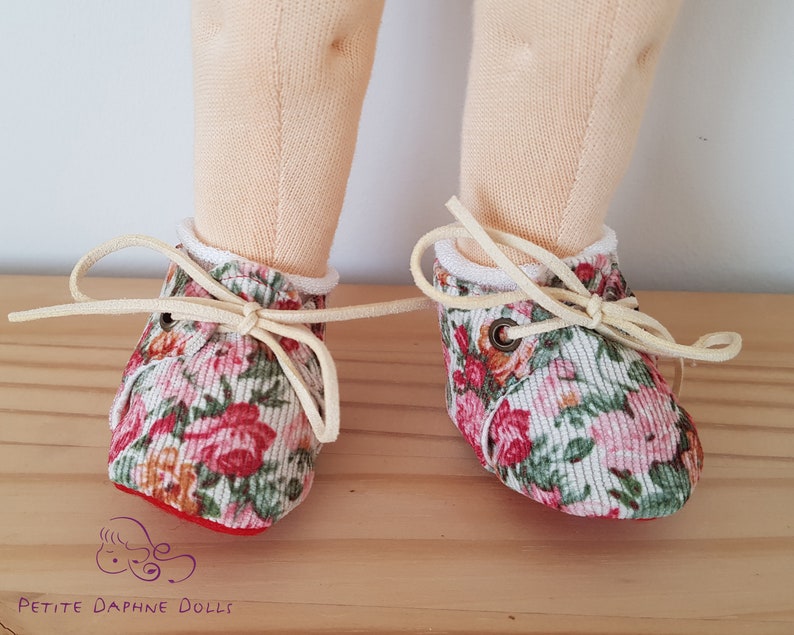 May include: A pair of floral print doll shoes with white laces and red trim. The shoes are tied with a bow and have a white trim around the top.