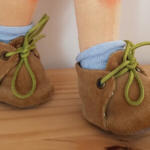 May include: A pair of brown corduroy baby shoes with green laces. The shoes have a light blue sock inside.