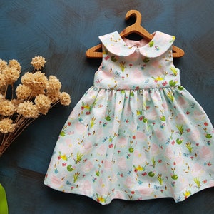 Puede incluir: Un vestido azul claro sin mangas con un cuello blanco de Peter Pan. El vestido tiene un estampado floral rosa y verde con ranas, mariposas y nen&uacute;fares.