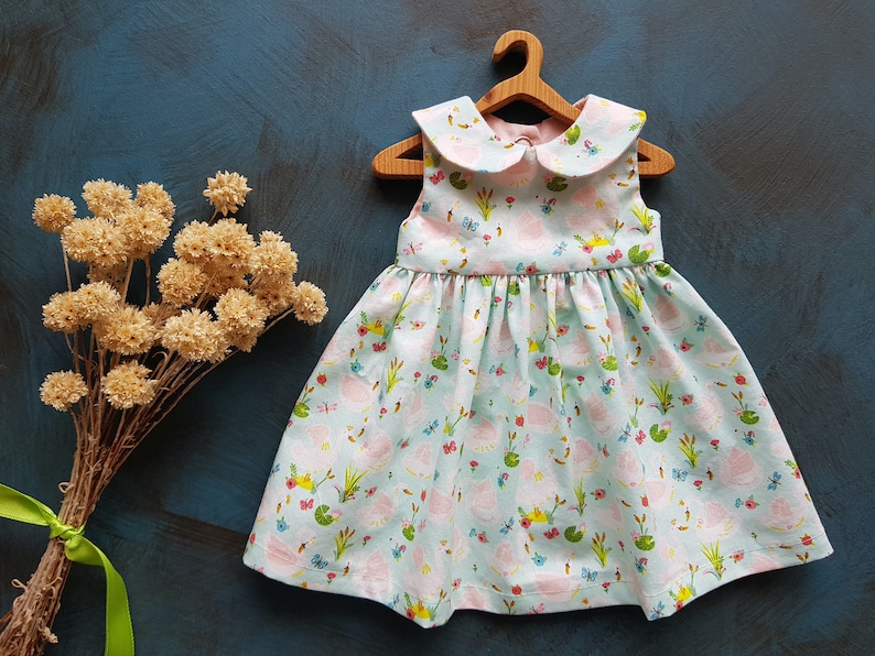 Puede incluir: Un vestido azul claro sin mangas con un cuello blanco de Peter Pan. El vestido tiene un estampado floral rosa y verde con ranas, mariposas y nen&uacute;fares.
