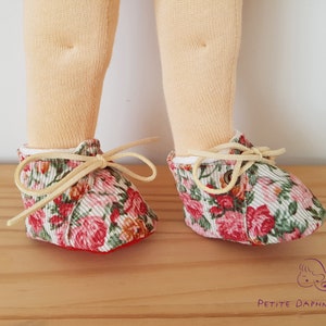 May include: A pair of doll shoes with a floral print. The shoes are made of a soft, pink and green fabric with a white background. The shoes have cream-colored laces and are tied in a bow. The shoes are sitting on a wooden surface.