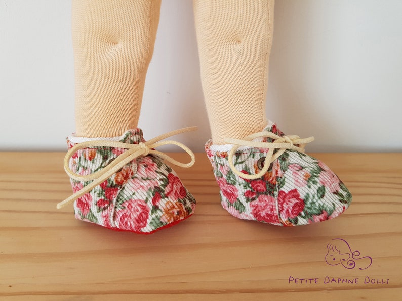May include: A pair of doll shoes with a floral print. The shoes are made of a soft, pink and green fabric with a white background. The shoes have cream-colored laces and are tied in a bow. The shoes are sitting on a wooden surface.