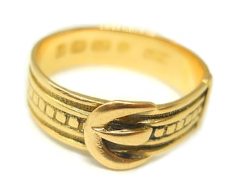 May include: A gold-colored ring with a buckle design. The ring features a detailed buckle clasp and decorative horizontal bands with small square accents. The ring is set against a white background.