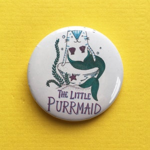 May include: A white button with a cartoon cat mermaid with a blue tail and purple seashells. The cat is sitting on a white seashell with a starfish. The text "The Little Purrmaid" is written in purple below the cat.