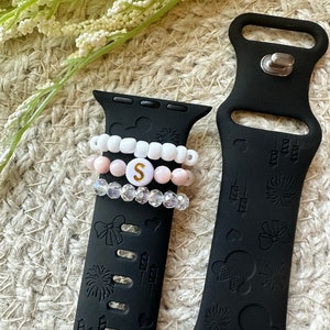 May include: Black Apple Watch band with a Mickey Mouse and fireworks design. The band has a white, pink, and clear beaded bracelet with a gold letter 'S' charm.