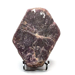 Op de afbeelding: Een ruwe, ongeslepen robijn edelsteen, met een diepe bordeauxrode kleur met lichtere roze en bruine insluitsels. De ruwe steen is gemonteerd op een kleine, zilverkleurige metalen displaystandaard, tegen een witte achtergrond. Het oppervlak van de robijn heeft een getextureerd, natuurlijk uiterlijk.