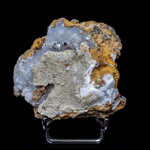 Op de afbeelding: Een close-up van een mineraalmonster met een mix van kleuren en texturen. De steen heeft een lichtgrijze, ruwe oppervlakte met vlekken van oranje-bruine en witte kristallijne formaties. Het monster wordt gepresenteerd op een metalen standaard tegen een zwarte achtergrond.