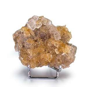 Puede incluir: Un grupo de cristales de color marrón dorado, posiblemente un espécimen mineral, se exhibe sobre un pequeño soporte de metal plateado. Los cristales tienen un aspecto rugoso y texturizado con tonos ámbar y beige. El fondo es blanco.