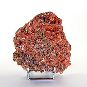 Könnte beinhalten: Ein lebhaftes rotes Mineralexemplar mit kristalliner Struktur. Das Mineral hat eine tiefe, satte rote Farbe mit einer rauen, strukturierten Oberfläche. Es wird auf einem kleinen, silbernen Metallständer präsentiert, der seine natürliche Schönheit hervorhebt.