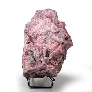 Puede incluir: Un espécimen mineral de rodocrosita en bruto, de color rosa intenso con zonas grises y negras. La roca se exhibe sobre un pequeño soporte metálico plateado sobre un fondo blanco. La textura del mineral es rugosa y cristalina.