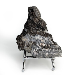 Può includere: Un esemplare minerale scuro e dalla consistenza ruvida con una formazione cristallina sulla sommità. La roccia è principalmente grigio scuro e nera, con sezioni grigie e bianche più chiare. È esposto su un piccolo supporto in metallo argentato.