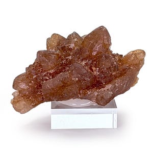 Könnte beinhalten: Ein Cluster aus bernsteinfarbenen Kristallen, die eine natürliche, raue Textur aufweisen. Die Kristalle sind auf einem transparenten, rechteckigen Sockel vor weißem Hintergrund montiert. Die Mineralformation hat eine komplexe, geschichtete Struktur mit Brauntönen.