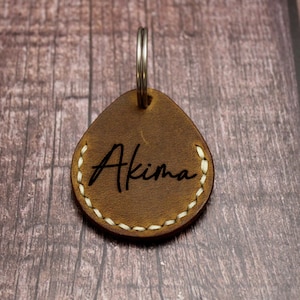 Può includere: Portachiavi in ​​cuoio marrone con un bordo cucito bianco e il nome "Akima" inciso in nero.