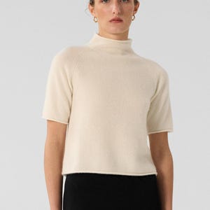 May include: A short-sleeved cream-colored mock neck top paired with a long black skirt. The top features a simple knit texture and a straight hemline, while the skirt is a solid black, ankle-length piece with a straight, streamlined silhouette.