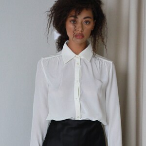 May include: A white button-down shirt with long sleeves and a classic collar. The shirt is paired with a black satin skirt. The outfit is simple and elegant, suitable for various occasions.