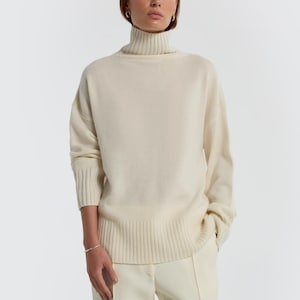 May include: A cream-colored turtleneck sweater and matching pants. The sweater has a ribbed hem and cuffs. The pants are a straight-leg style. The outfit is a neutral, minimalist ensemble.