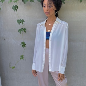 May include: A cream-colored button-down shirt paired with blush-colored wide-leg pants. The outfit includes a blue and white patterned top and nude-colored square-toe sandals. A gold necklace adds a touch of elegance.