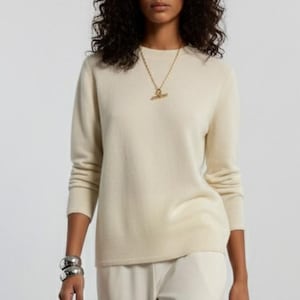 May include: A cream-colored long-sleeved sweater paired with matching cream-colored wide-leg trousers. The outfit is accessorized with a gold-toned chain necklace featuring a T-bar pendant and a thick silver-toned cuff bracelet on the wrist.