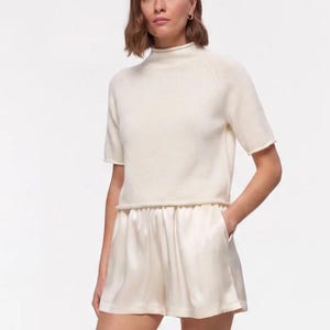 May include: A cream-colored short-sleeved mock neck sweater with a raw hem paired with cream-colored satin shorts featuring an elasticized waistband and side pockets, worn with matching cream-colored open-toe slide sandals.