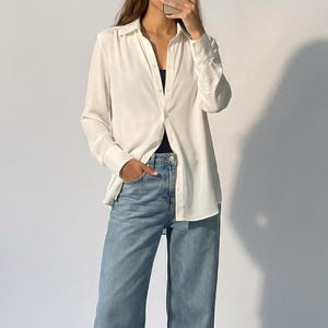 May include: A white button-down shirt with long sleeves and light blue jeans. The shirt is partially unbuttoned, revealing a dark top underneath. The jeans have a wide leg design. Black shoes complete the outfit.