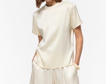 Puede incluir: Una camiseta de manga corta de color crema con cuello redondo y pantalones cortos a juego del mismo color. Ambas prendas están confeccionadas con un tejido liso y lustroso con un brillo sutil. La parte superior tiene un dobladillo recto.