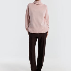 May include: A light pink turtleneck sweater paired with dark brown trousers. The sweater has long sleeves and a ribbed hem. The trousers are straight-legged and have a front seam. The outfit is completed with black sandals.