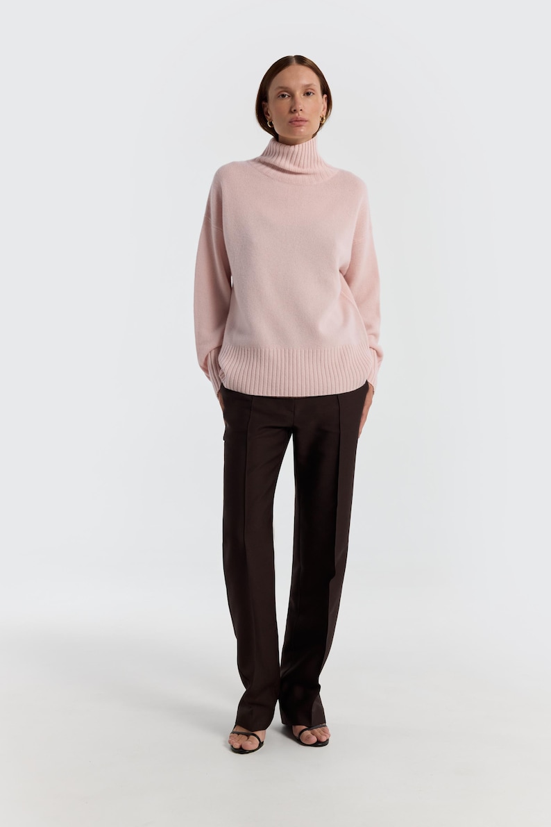 May include: A light pink turtleneck sweater paired with dark brown trousers. The sweater has long sleeves and a ribbed hem. The trousers are straight-legged and have a front seam. The outfit is completed with black sandals.