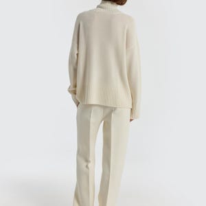 May include: A cream-colored turtleneck sweater and matching pants. The sweater has a ribbed hem and cuffs. The pants are wide-legged. The outfit is made of a soft, comfortable material. The model is facing away from the camera.