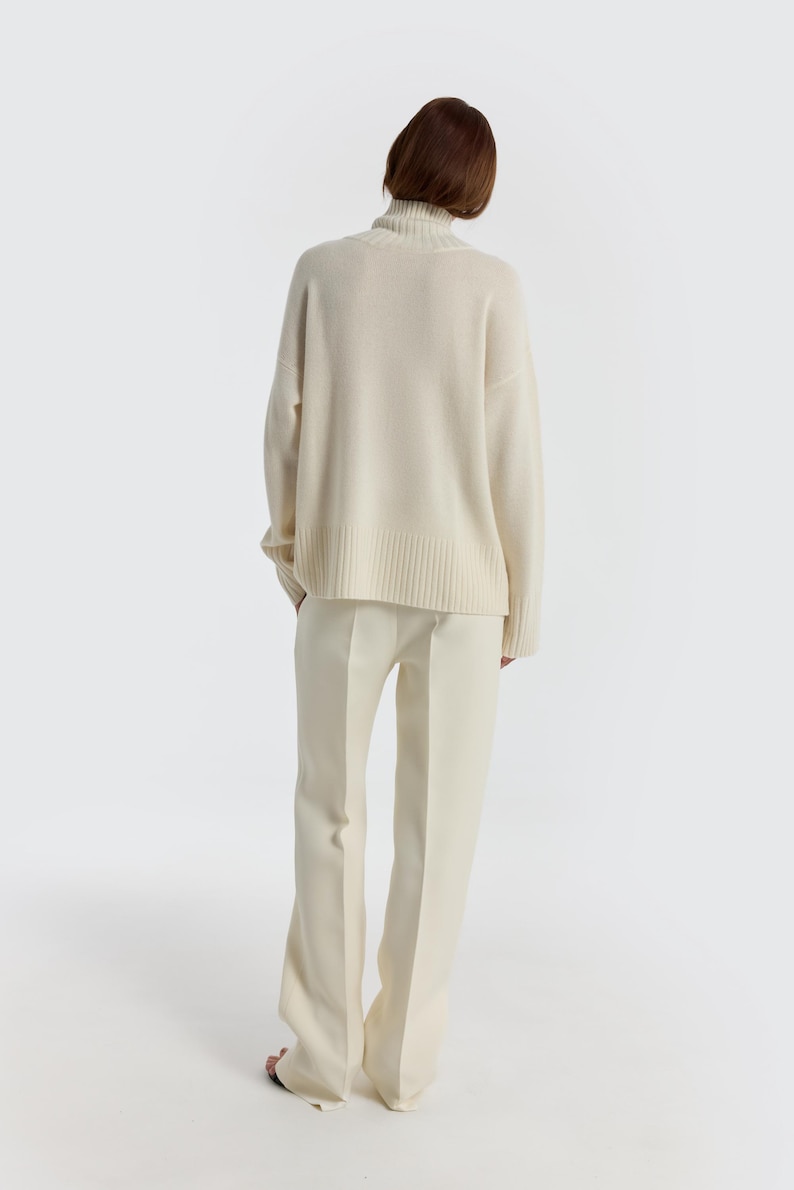 May include: A cream-colored turtleneck sweater and matching pants. The sweater has a ribbed hem and cuffs. The pants are wide-legged. The outfit is made of a soft, comfortable material. The model is facing away from the camera.