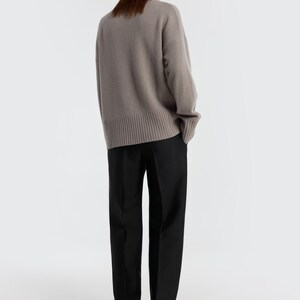 May include: A person wearing a taupe-colored sweater and black pants. The sweater has a ribbed hem and cuffs. The pants are straight-legged. The background is plain white. The clothing is likely part of a fall or winter collection.