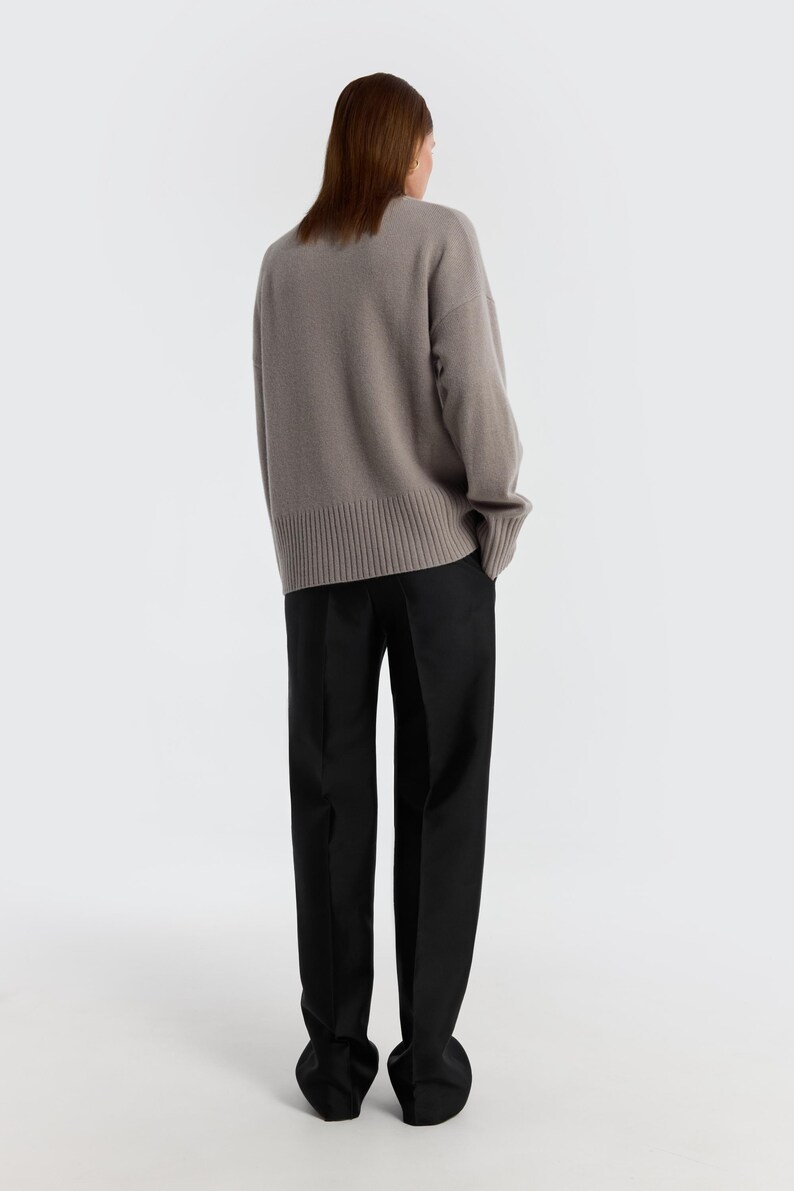 May include: A person wearing a taupe-colored sweater and black pants. The sweater has a ribbed hem and cuffs. The pants are straight-legged. The background is plain white. The clothing is likely part of a fall or winter collection.