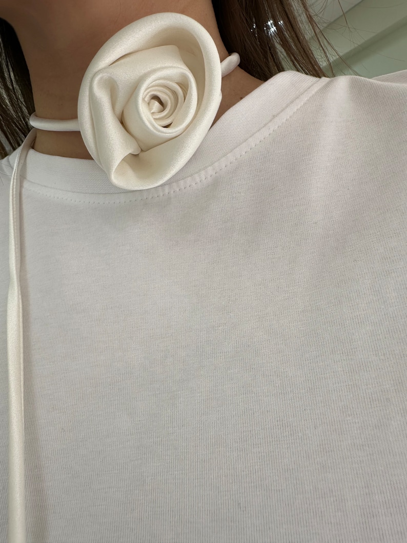 May include: A white satin rose choker necklace. The rose is made of fabric and is tied together with a white satin ribbon.