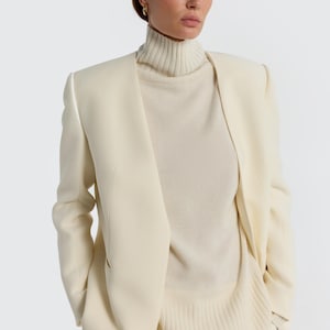 May include: A cream-colored blazer with a matching turtleneck sweater and pants. The blazer has a tailored fit with a notched lapel and long sleeves. The turtleneck sweater is a lighter shade of cream. The outfit is a monochromatic ensemble.