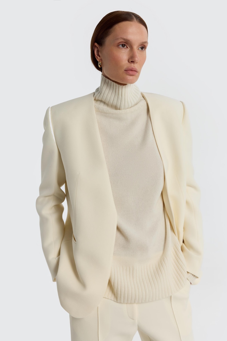 May include: A cream-colored blazer with a matching turtleneck sweater and pants. The blazer has a tailored fit with a notched lapel and long sleeves. The turtleneck sweater is a lighter shade of cream. The outfit is a monochromatic ensemble.