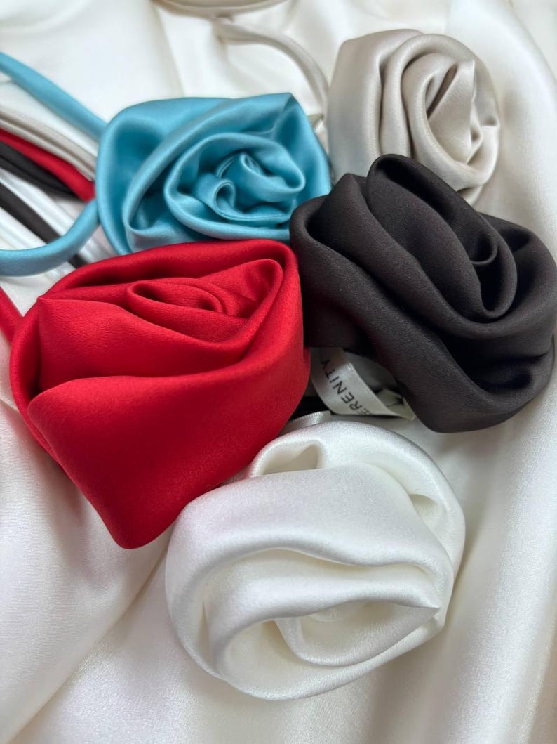 May include: Five silk rose hair ties in different colors: white, light blue, red, black, and cream. The hair ties are arranged in a circle on a white background.