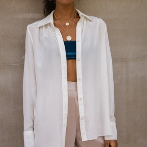 May include: A woman wearing a white button-down shirt, layered gold necklaces, and blush-colored satin pants. A teal bandeau top is visible beneath the open shirt. The outfit is styled against a neutral background.