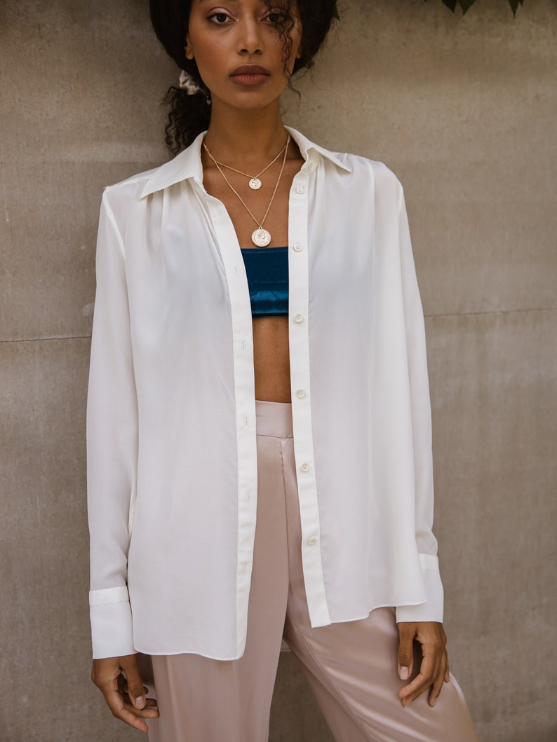 May include: A woman wearing a white button-down shirt, layered gold necklaces, and blush-colored satin pants. A teal bandeau top is visible beneath the open shirt. The outfit is styled against a neutral background.