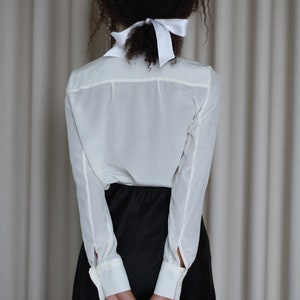 May include: A person wearing a white button-down shirt with long sleeves and a black skirt, holding a black briefcase. A white bow is tied in the person's hair. The background is a neutral-colored curtain.