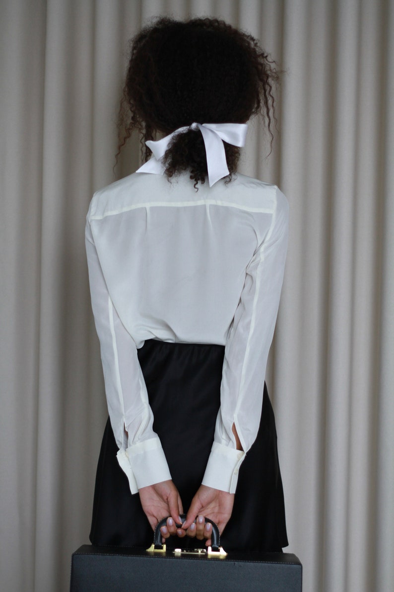 May include: A person wearing a white button-down shirt with long sleeves and a black skirt, holding a black briefcase. A white bow is tied in the person's hair. The background is a neutral-colored curtain.