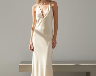 Op de afbeelding: Een crèmekleurige, vloerlange slipdress met een diepe V-hals en spaghettibandjes. De jurk is gemaakt van een soepele, vloeiende stof en heeft een eenvoudig, elegant ontwerp. De jurk is geschikt voor formele evenementen.
