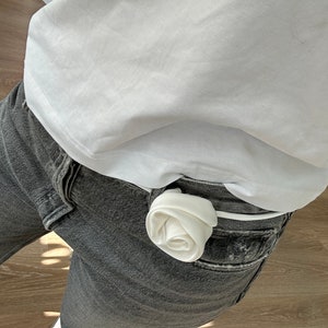 May include: A white fabric rose tucked into the waistband of a pair of gray jeans.