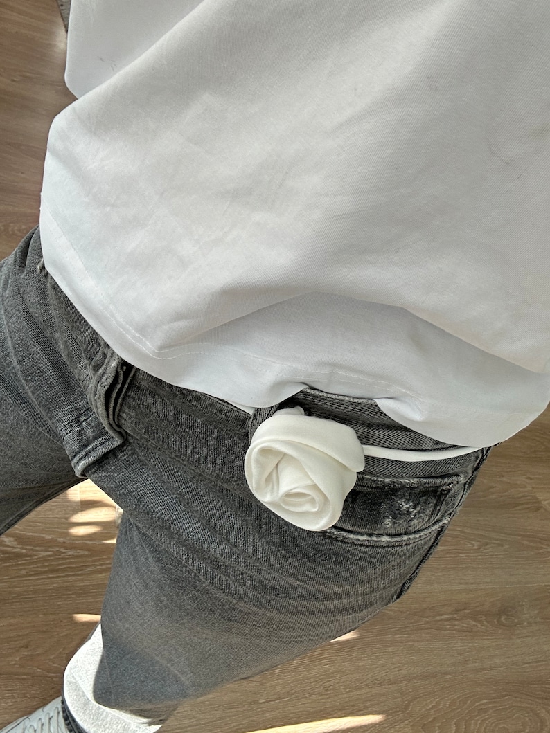May include: A white fabric rose tucked into the waistband of a pair of gray jeans.