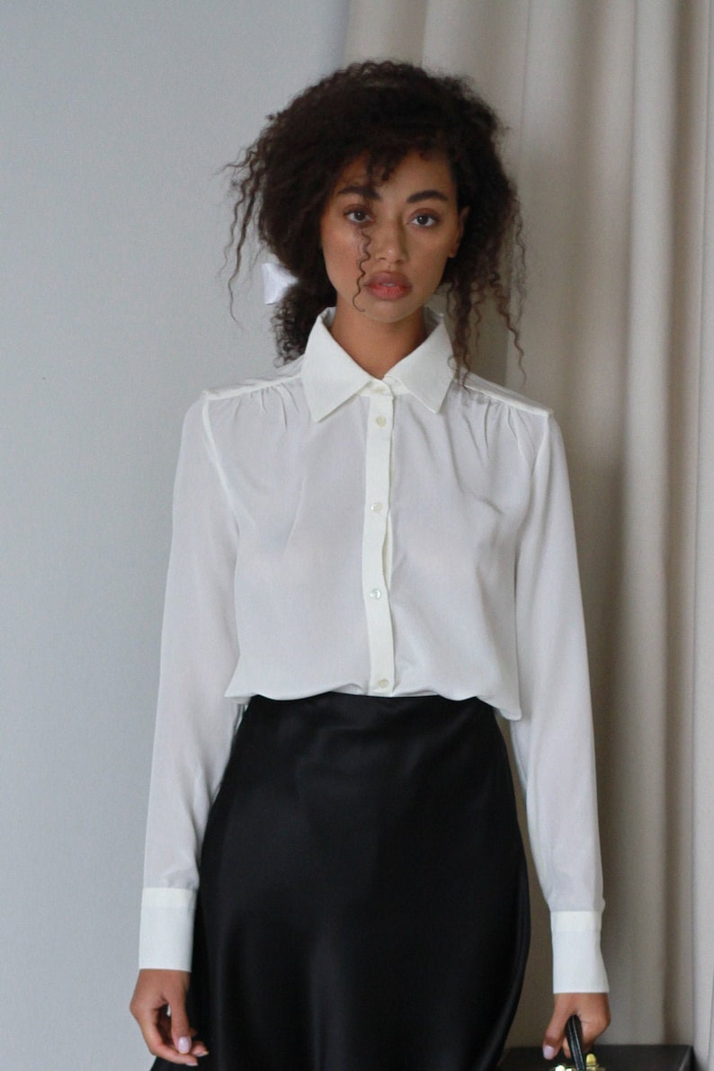 May include: A white button-down shirt with long sleeves and a classic collar. The shirt is paired with a black satin skirt. The outfit is simple and elegant, suitable for various occasions.
