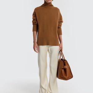 May include: A woman wearing a brown turtleneck sweater and cream-colored pants. She is holding a brown leather tote bag. The outfit is simple and elegant, with a focus on neutral tones and clean lines.