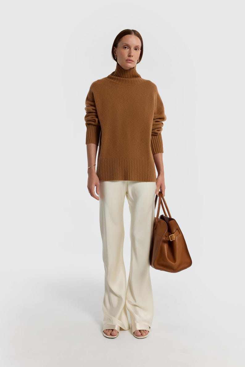 May include: A woman wearing a brown turtleneck sweater and cream-colored pants. She is holding a brown leather tote bag. The outfit is simple and elegant, with a focus on neutral tones and clean lines.