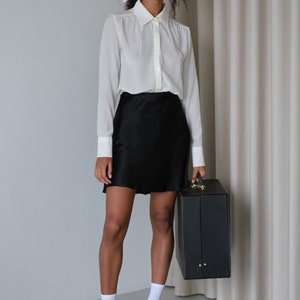 May include: A woman wearing a white button-down shirt with a black skirt and black loafers. She is holding a black briefcase. The outfit is simple and elegant.